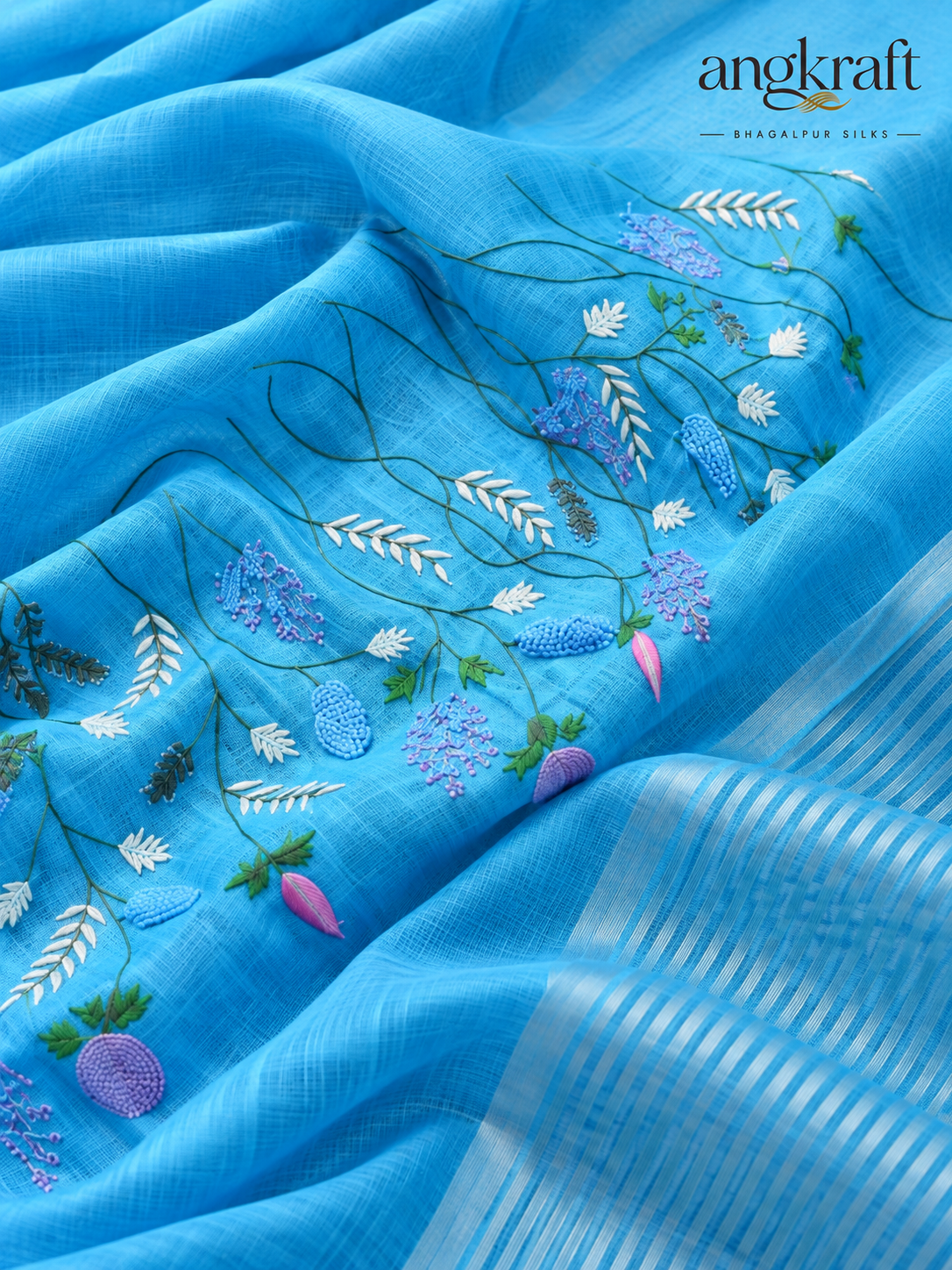 A close-up of a light blue linen saree fabric with detailed floral and botanical embroidery in shades of purple, white, and green, featuring a striped border on the right side and the 'angkraft' brand logo in the top right corner.