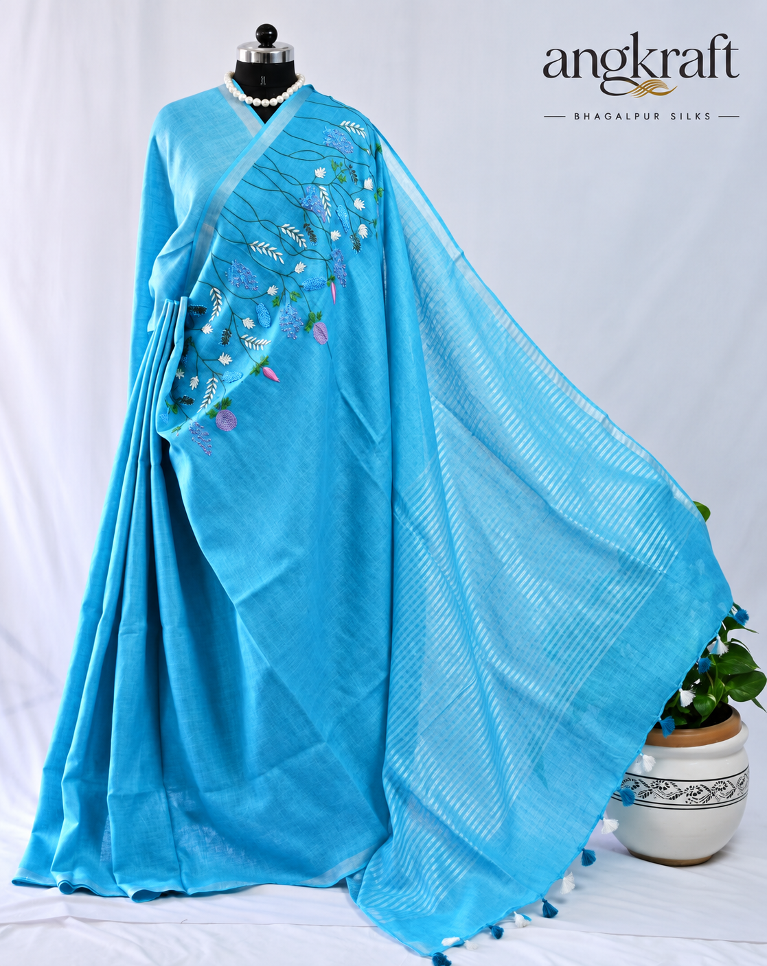 A bright blue linen saree draped on a mannequin, featuring delicate floral embroidery on the border and a matching blue patterned pallu. The brand logo 'angkraft' is visible in the top right corner.