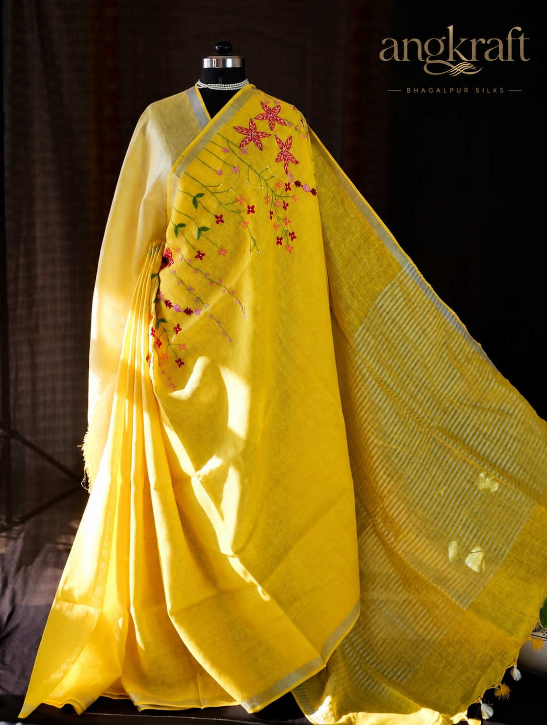 A bright yellow Linen saree with pink and green floral embroidery and a striped pallu, displayed on a black mannequin against a dark background. The Angkraft brand logo is visible in the top right corner.